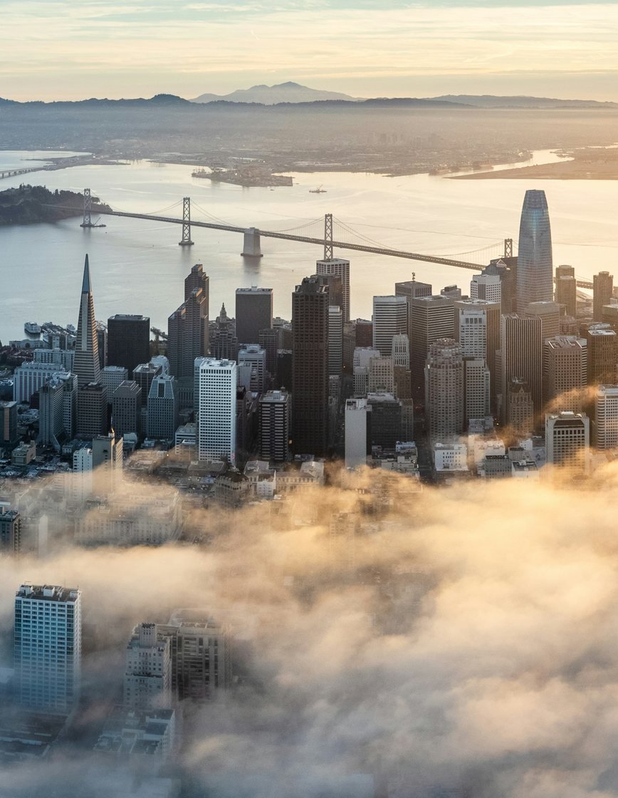 Aerial view of downtown San Francisco at golden hour, looking across the financial district toward the bay and the Bay Bridge